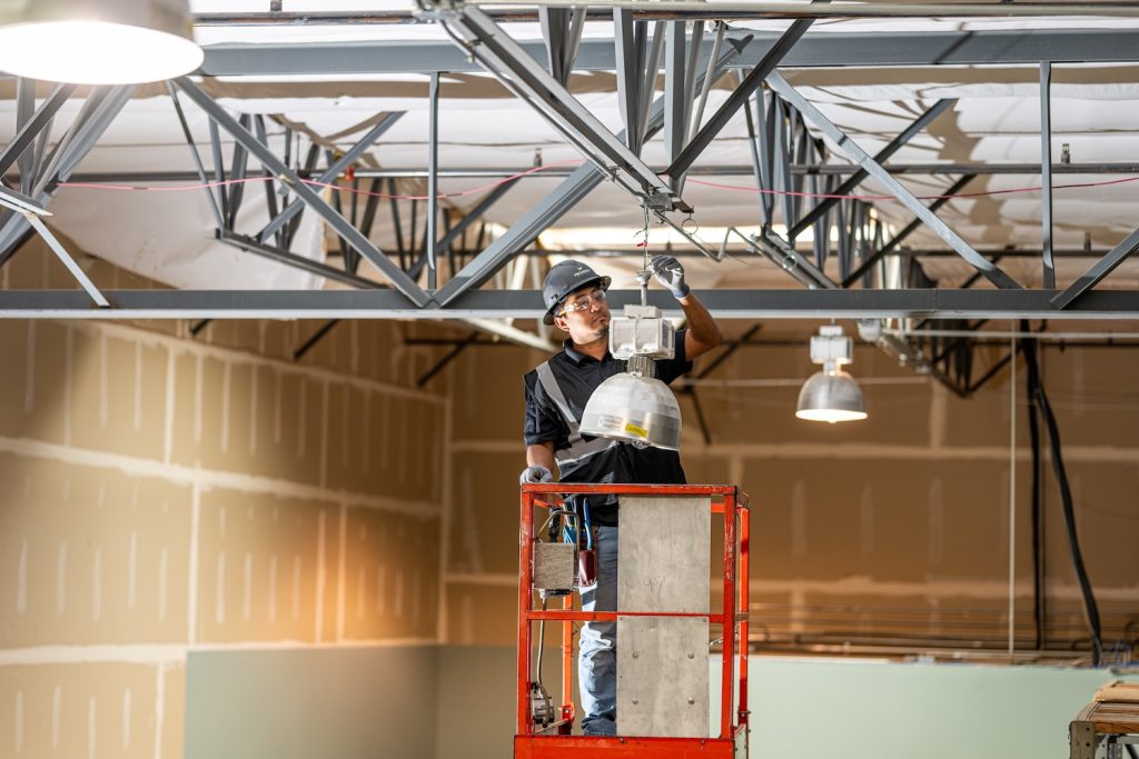 TCE_October blog image | TechCity Electric Construction worker on a red lift installing a ceiling light fixture in an unfinished interior. Safety gear includes a hard hat and gloves.